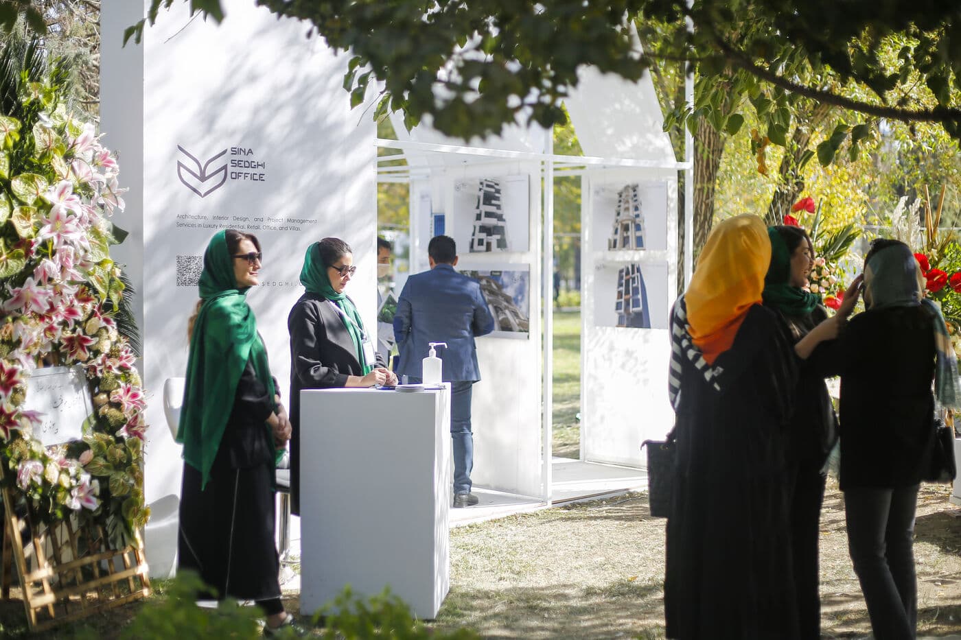 بازدیدکنندگان کنار یک غرفهی سفید در سایهروشن درختان، در میانهی گفتوگو با مشارکتکننده
